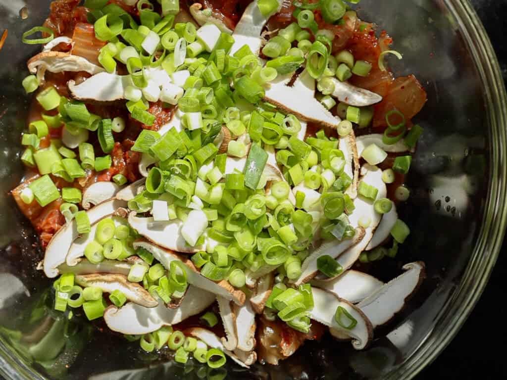 A glass bowl filled with chopped green onions, sliced mushrooms, and kimchi, viewed from above. Bright sunlight highlights the fresh ingredients, emphasizing their colors and textures.